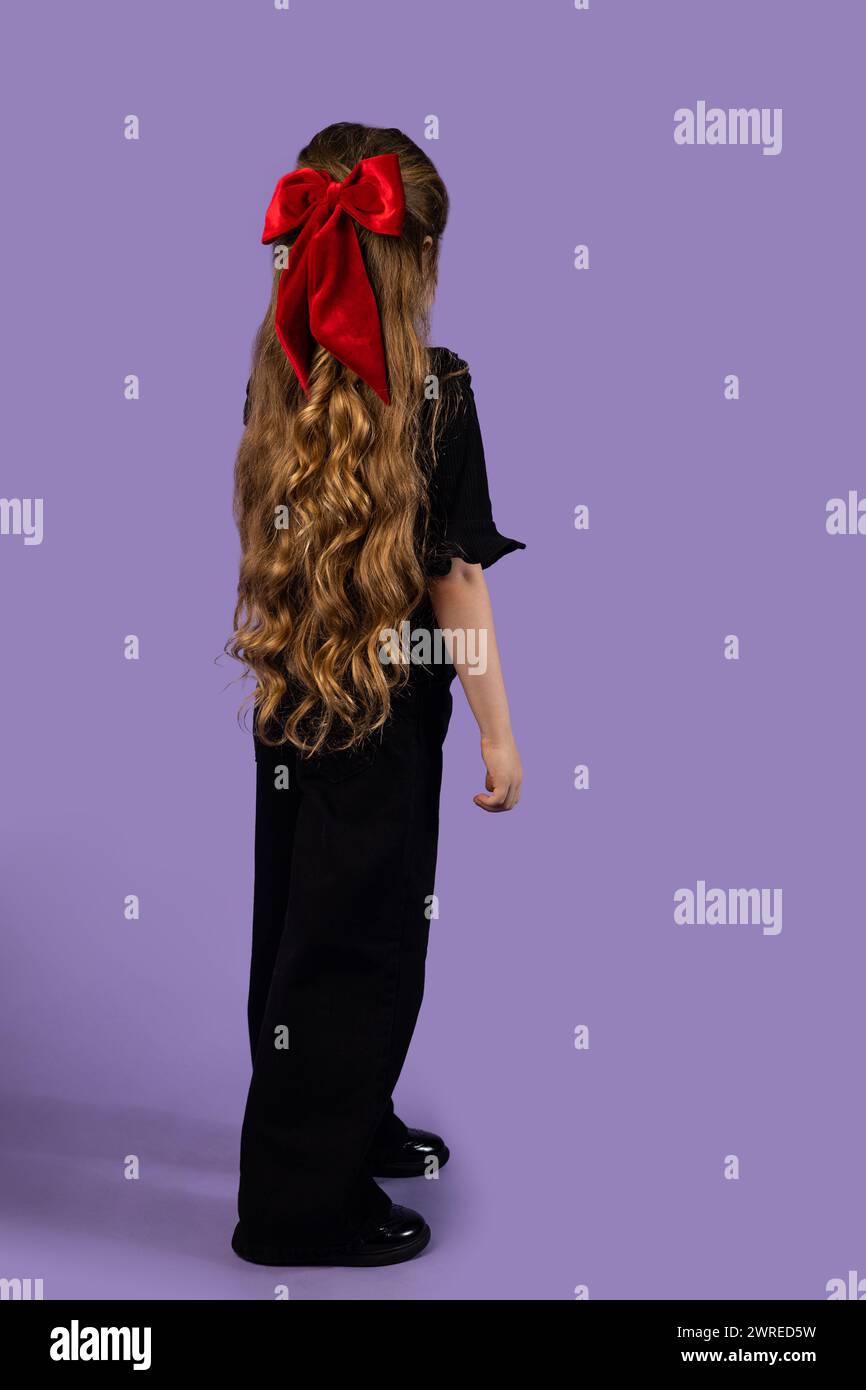 Ripresa posteriore di una bambina con lunghi capelli biondi ondulati vestiti di nero e un fiocco rosso nei capelli che le guardano dietro, girata su uno studio viola Foto Stock