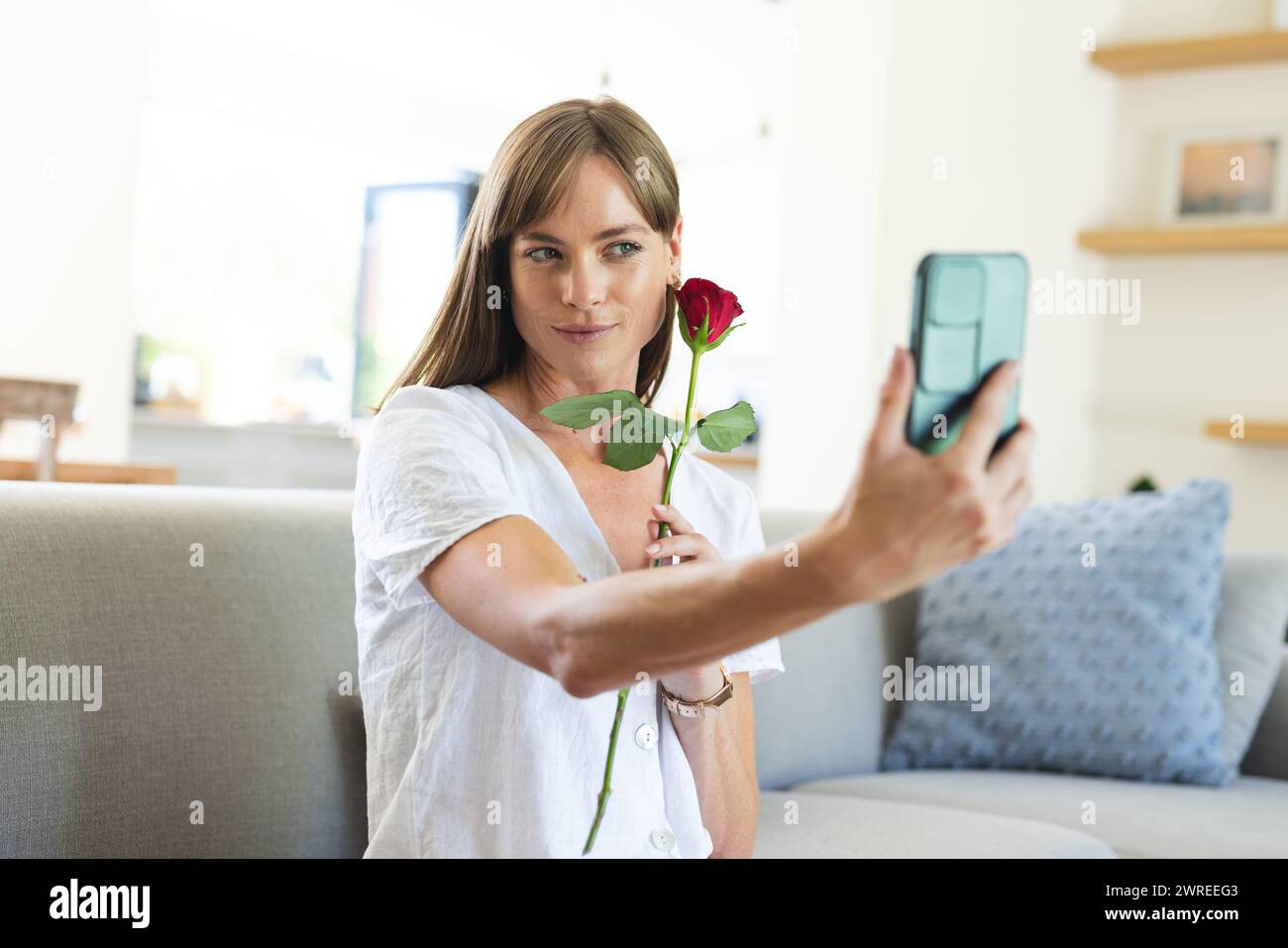 Una donna caucasica fa un selfie con una rosa rossa, sorridendo dolcemente al suo telefono Foto Stock