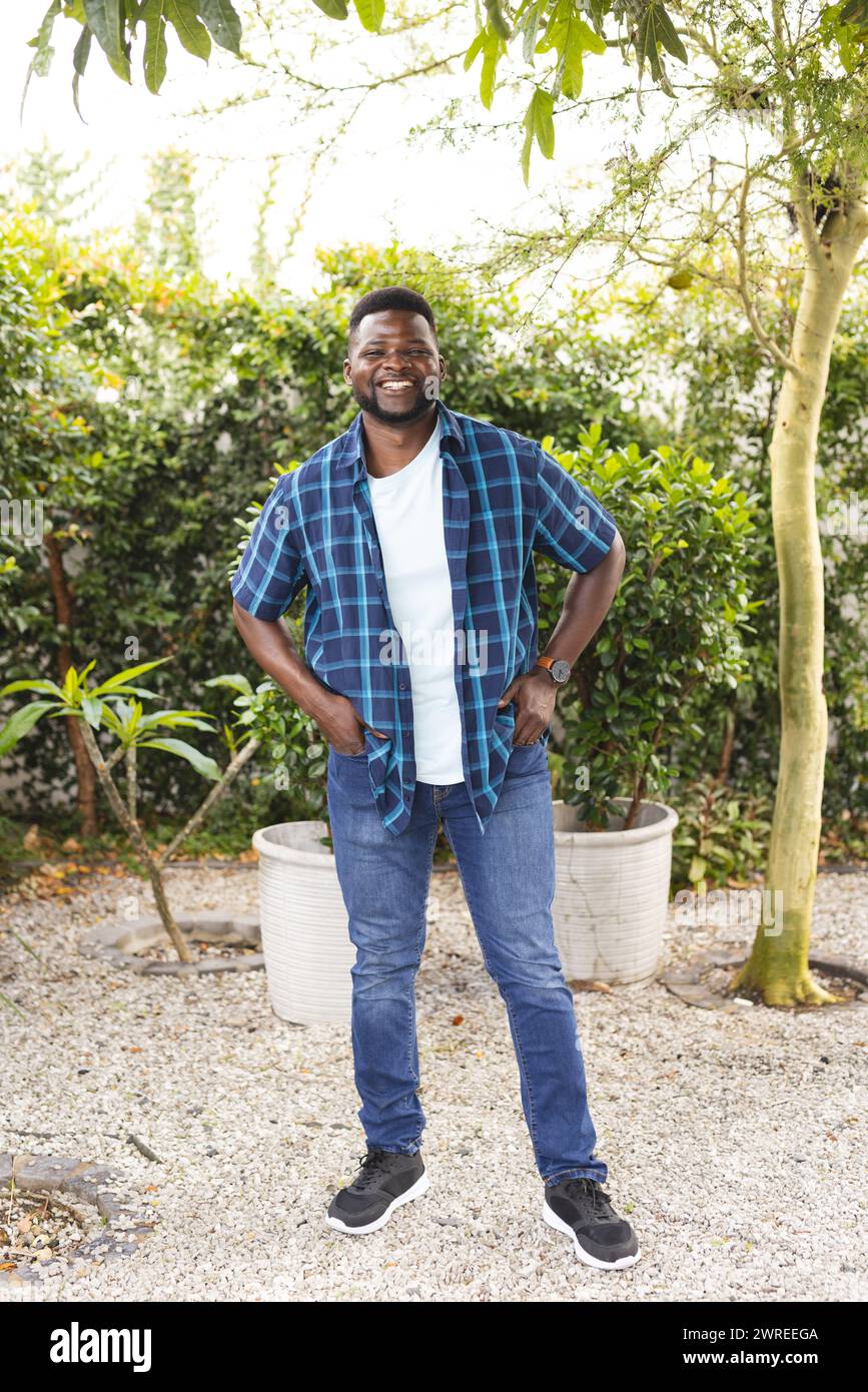 Un uomo afroamericano sorride in un giardino, indossando una camicia a quadri blu e jeans Foto Stock