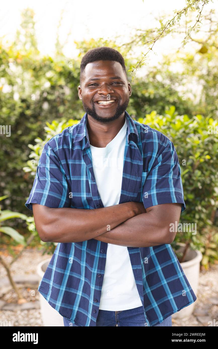 L'uomo afroamericano sorride in grande, indossa una camicia blu a quadri e sta in piedi all'aperto in un giardino Foto Stock