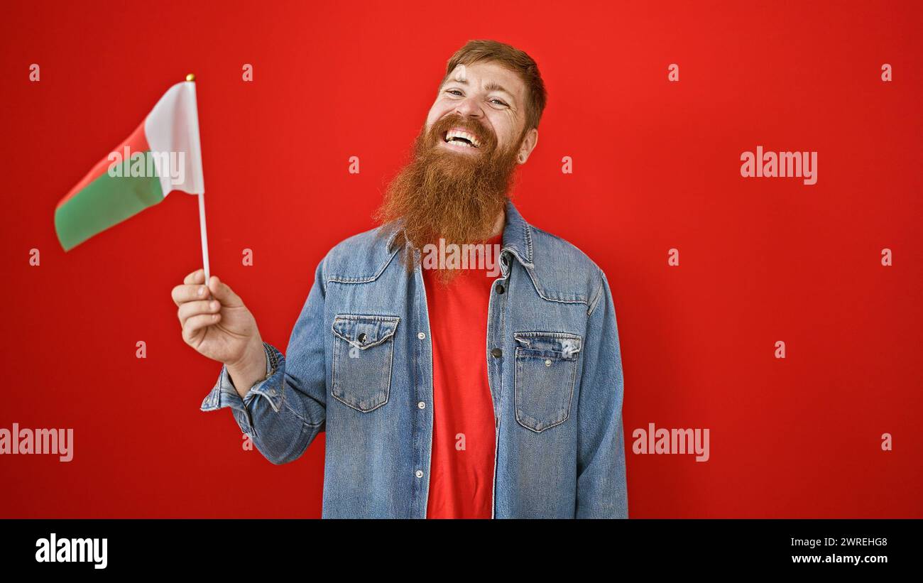 Felice giovane rossa, pieno di fiducia e gioia, in piedi casualmente, sventolando la bandiera del madagascar sullo sfondo di un muro rosso isolato. suo Foto Stock