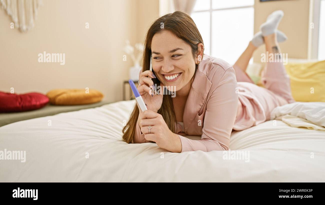 Una donna gioiosa giace sul letto nella sua camera da letto, tenendo un test di gravidanza e parlando al telefono. Foto Stock