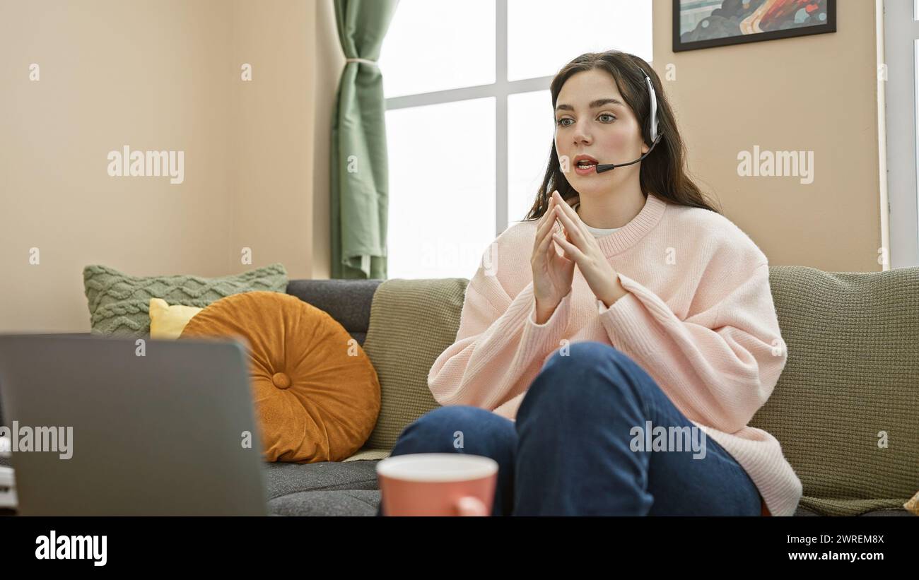 Una giovane donna che indossa una cuffia parla tranquillamente in casa, magari coinvolta in un telelavoro, in un ambiente informale da ufficio domestico in salotto. Foto Stock