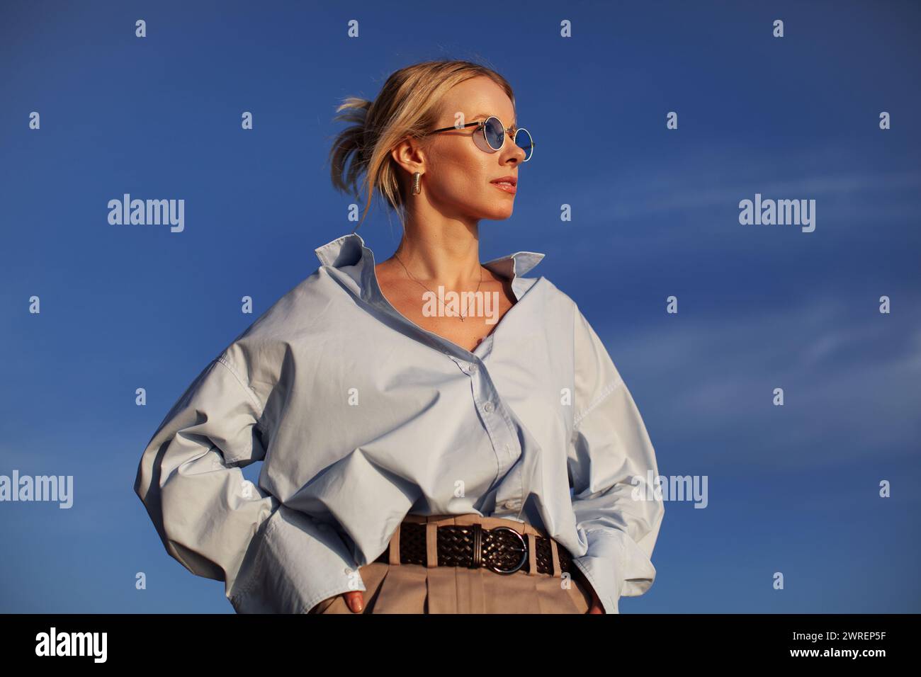 Donna moderna con una camicia oversize e occhiali da sole si staglia all'aperto contro il cielo. Chic, contemporaneo ed elegante, cattura la libertà estiva. Foto Stock