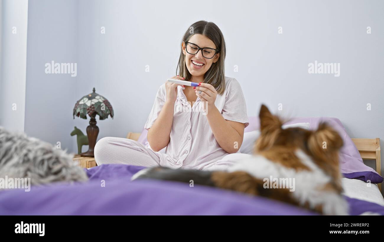La giovane donna eccitata legge un test di gravidanza positivo in camera da letto con cani nelle vicinanze. Foto Stock