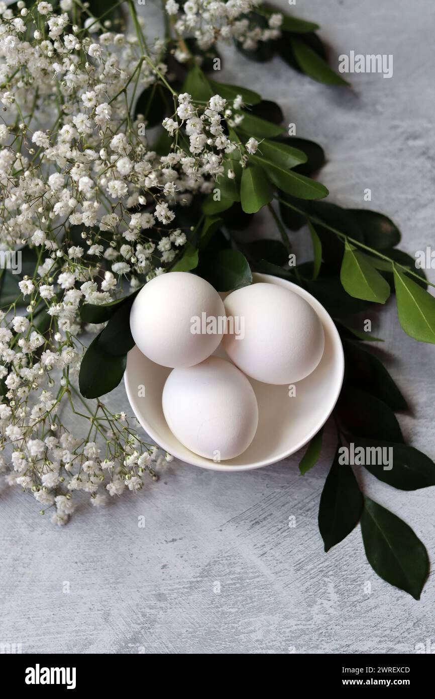 Composizione semplice natura morta con uova e fiori bianchi. Concetto di festa di Pasqua. Uova in un recipiente di ceramica bianco, foto vista dall'alto. Foto Stock