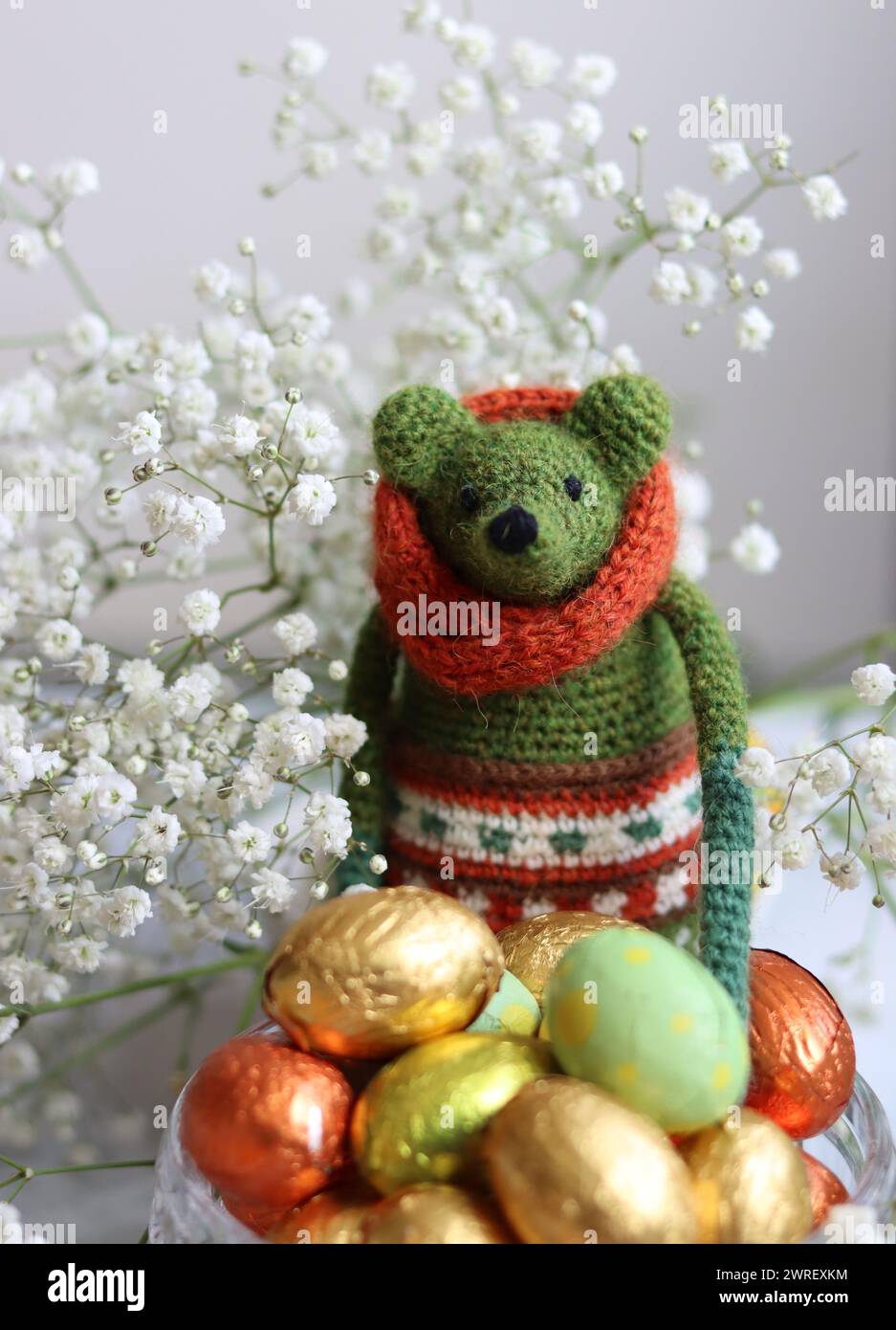 Giocattolo verde all'uncinetto, uova di cioccolato e fiori bianchi su un tavolo. Regalo fatto a mano per Pasqua. Foto colorate di natura morta con dolci avvolti in olio. Foto Stock