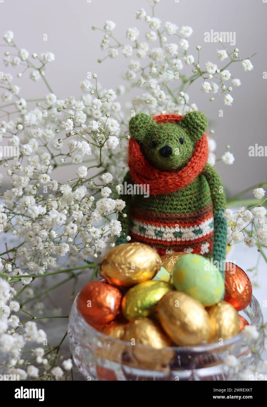 Giocattolo verde all'uncinetto, uova di cioccolato e fiori bianchi su un tavolo. Regalo fatto a mano per Pasqua. Foto colorate di natura morta con dolci avvolti in olio. Foto Stock