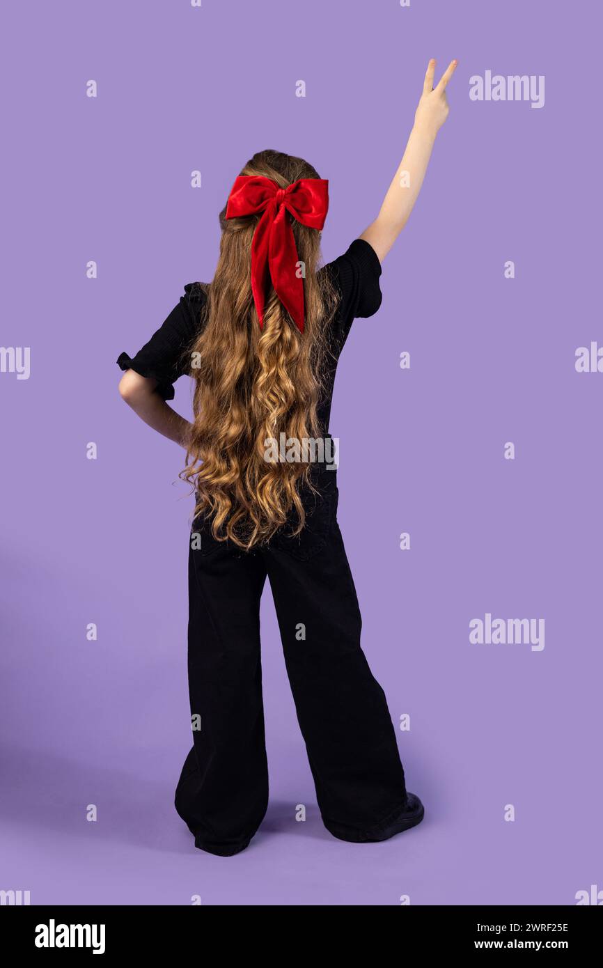 Foto a tutta lunghezza della bambina bionda che mostra un cartello a V con t-shirt, jeans e sneakers nere isolate su uno sfondo viola pastello chiaro. Alto Foto Stock