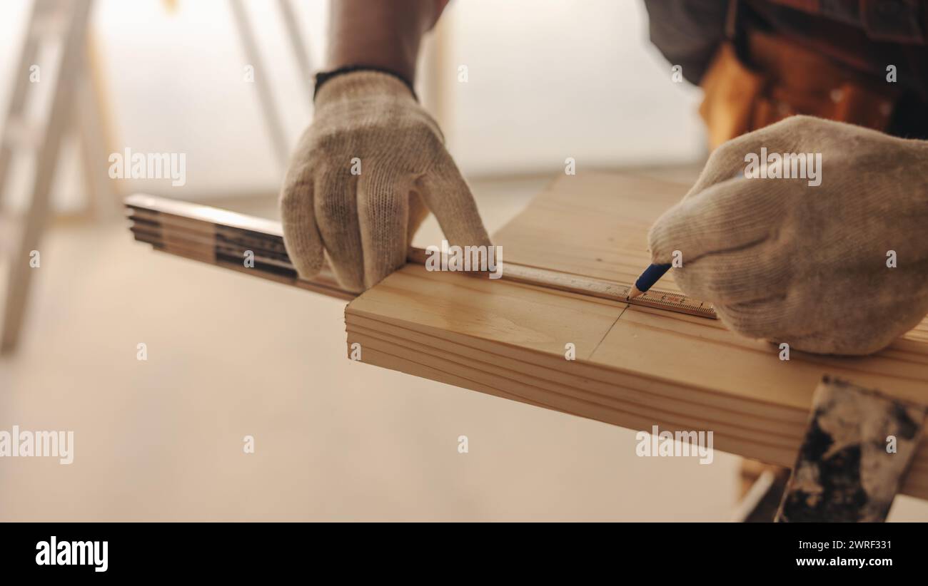Il carpentiere maschio nei guanti da lavoro misura e segna una tavola di legno all'interno di una casa. Questo lavoratore esperto offre competenze per migliorare gli interni della casa. H Foto Stock