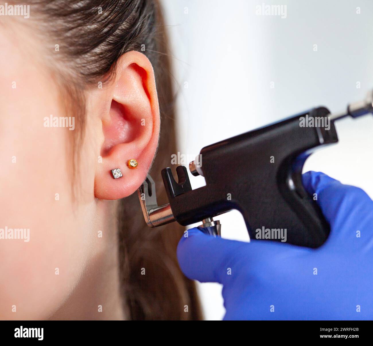 Un medico in guanti medici blu sterili perfora le orecchie di una giovane ragazza nello studio medico con una pistola perforante nera. Foto di alta qualità Foto Stock