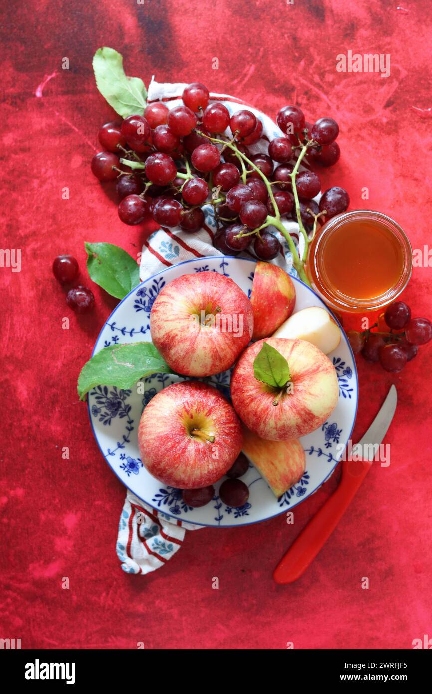 Mele rosse e uva su fondo rosso con una tazza di miele. Festa ebraica di Capodanno Rosh Hashana. Natura morta con frutta di stagione e miele. Foto Stock