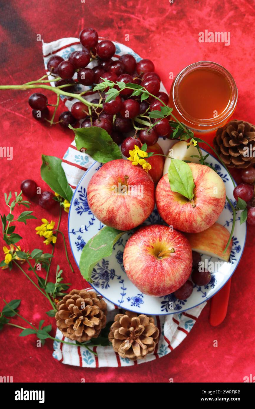 Mele rosse e uva su fondo rosso con una tazza di miele. Festa ebraica di Capodanno Rosh Hashana. Natura morta con frutta di stagione e miele. Foto Stock