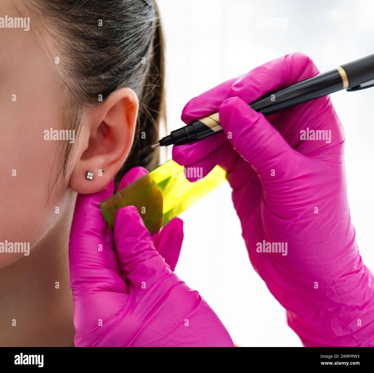Le mani di una cosmetologa indossate con guanti sterili rosa tengono in mano un righello e una penna per misurare la posizione dell'orecchio di una paziente e perforare l'orecchino. Alta qualità p Foto Stock