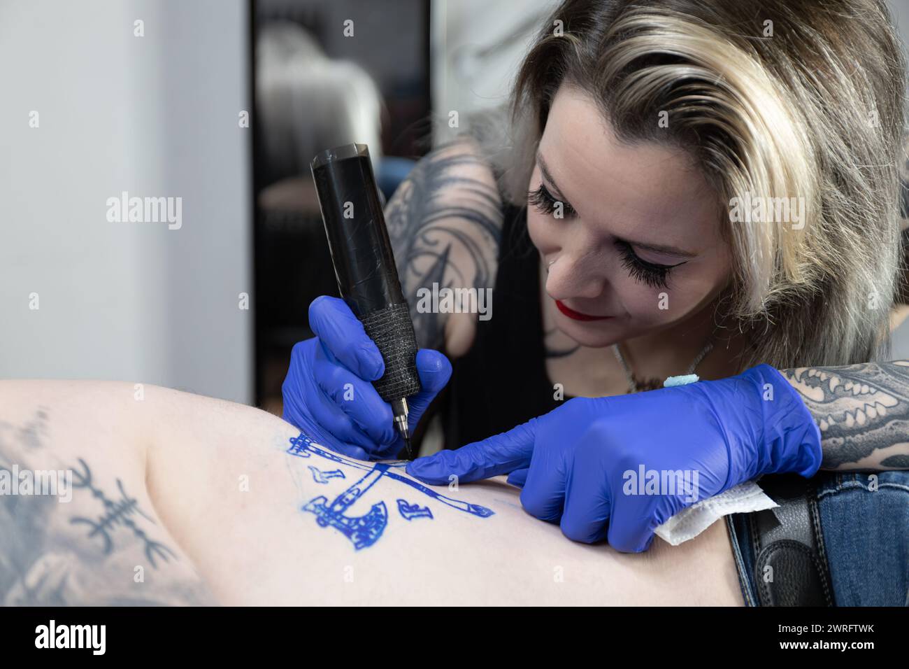 Foto orizzontale messa a fuoco intensa mentre un tatuatore tatua uno stemma sul corpo di un cliente, dando vita al design. Concetto di business, arte. Foto Stock