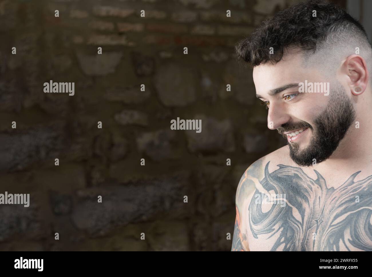 Foto orizzontale un giovane con un ampio sorriso mostra il suo intricato tatuaggio sul petto, trasudando sicurezza e stile su uno sfondo rustico. Copia s Foto Stock