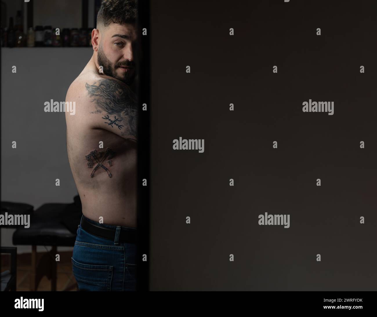 Foto orizzontale un uomo tatuato guarda indietro, la sua body art visibile in uno studio di tatuaggio sereno e poco illuminato, che evoca una narrazione di espressione personale. Poliziotto Foto Stock