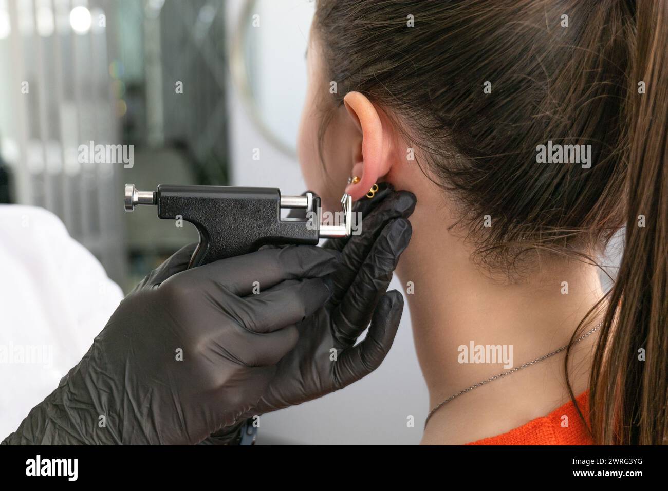 Un medico in guanti medici neri perfora le orecchie di una bella giovane donna nello studio medico utilizzando una pistola piercing nera sicura e orecchino medico Foto Stock