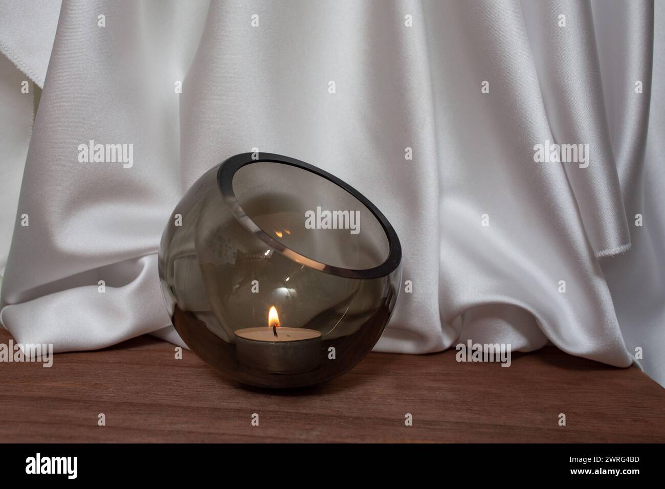 Ciotola di vetro con candela accesa su pavimento in legno con eleganti tende in tessuto satinato bianco ondulato, sfocata sullo sfondo, sfondo per il posizionamento dei prodotti Foto Stock