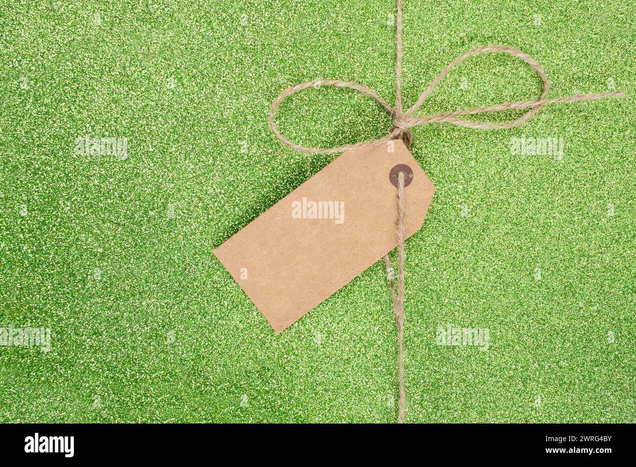 Filetto di iuta con un semplice arco e un'etichetta vuota di cartone marrone su sfondo verde glitter Foto Stock