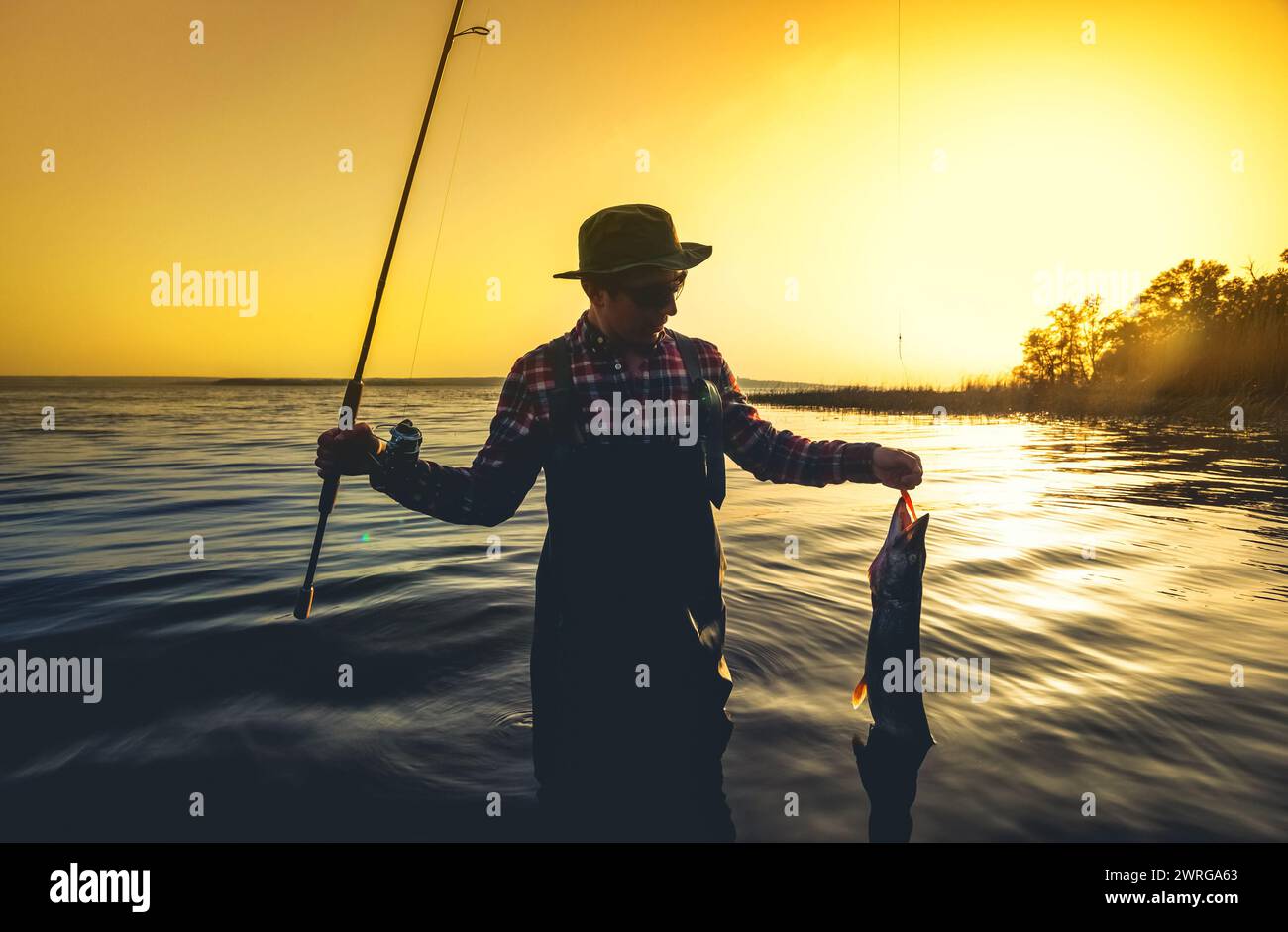 Un pescatore con una canna da pesca in mano e un pesce catturato si staglia in acqua davanti a un bellissimo tramonto. Foto Stock