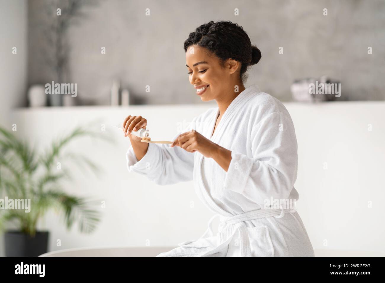 Felice donna nera in accappatoio che si prepara a lavarsi i denti, applicando dentifricio Foto Stock