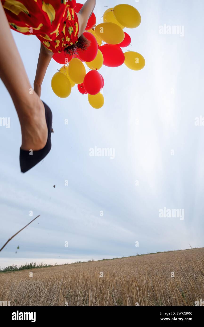 sparato dal basso di una donna bruna con palloncini gialli e rossi che corrono e saltano all'aperto. I colori rosso e giallo dominano. Foto Stock