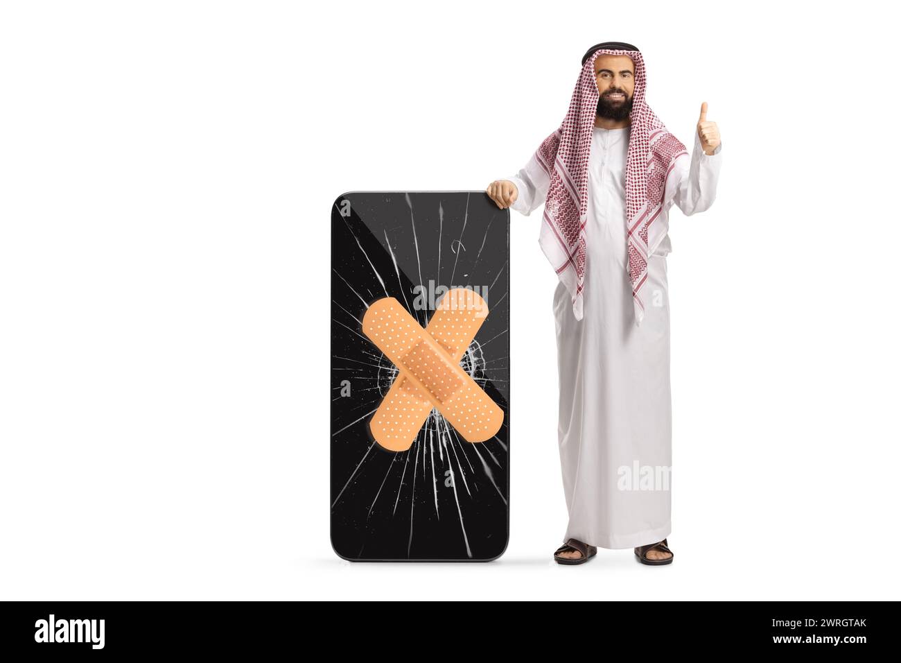 Uomo arabo saudita in abiti etnici che gestiva i pollici in alto e in piedi accanto a un telefono cellulare con schermo incrinato isolato su sfondo bianco Foto Stock