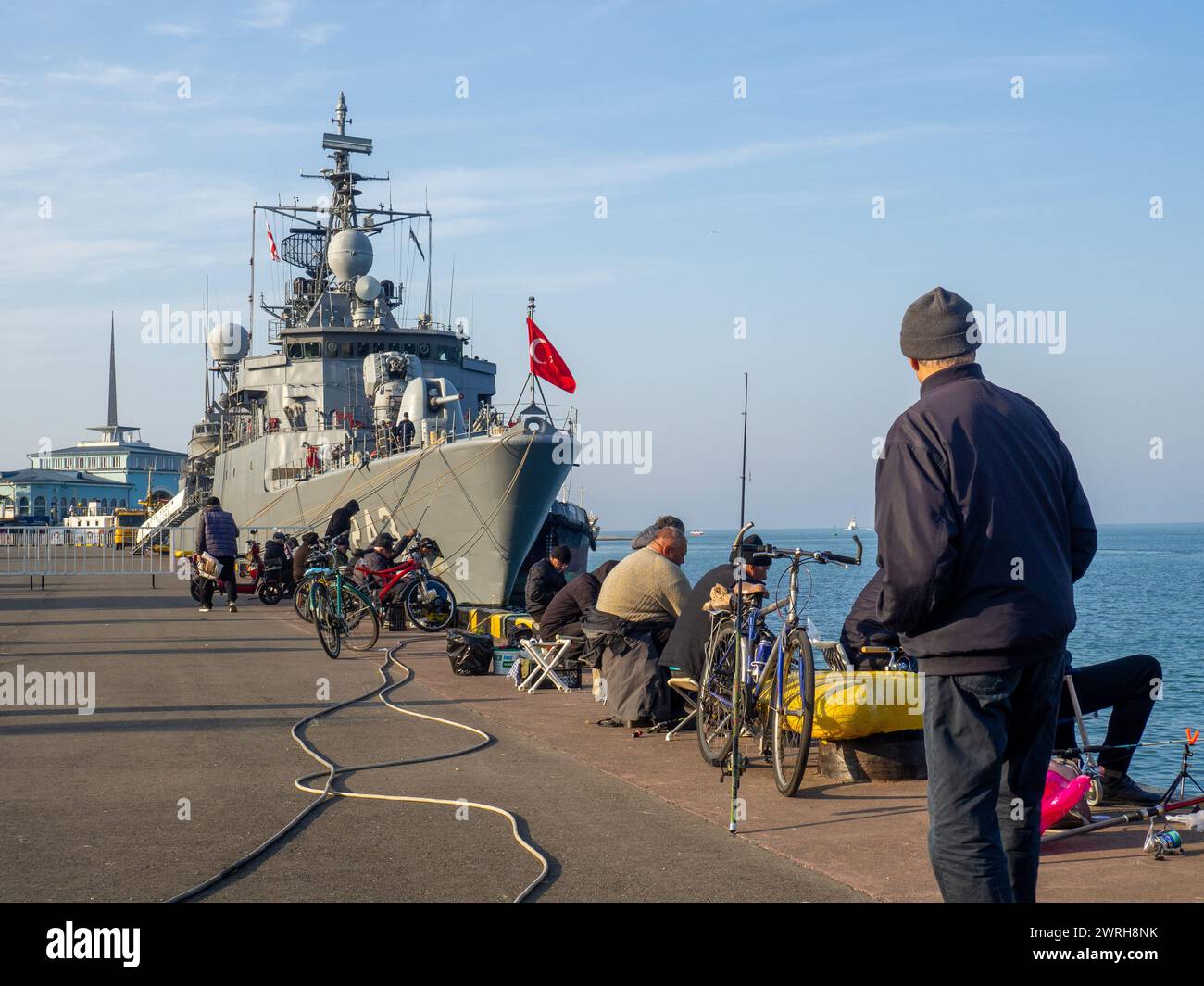 Batumi, Georgia. 03.05.2024 residenti locali che praticano la pesca in mare amatoriale. I pescatori catturano il pesce con le canne da pesca accanto a una nave da guerra. Una nave da guerra è ormeggiata Foto Stock