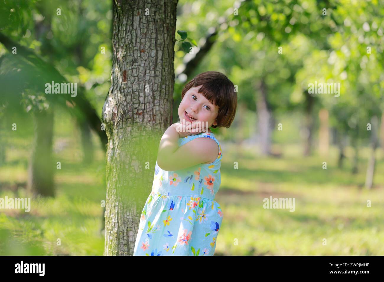 Una bambina carina che gioca sotto gli alberi da frutto, vestita con abiti estivi. Alberi verdi sfocati sullo sfondo. Giocare sotto gli alberi da frutto e mangiare frutta Foto Stock