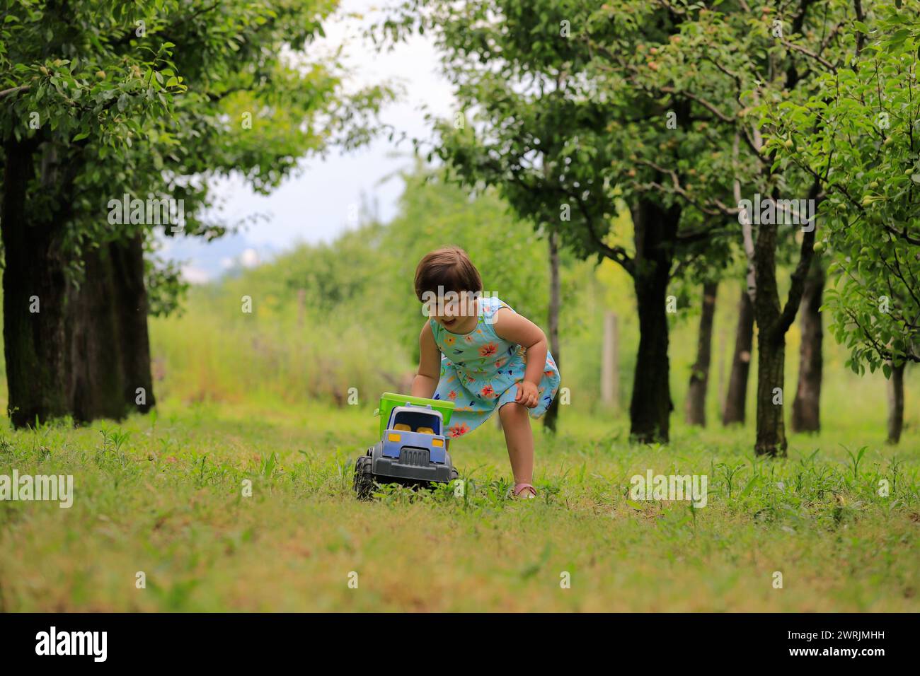 Una bambina carina che gioca sotto gli alberi da frutto, vestita con abiti estivi. Alberi verdi sfocati sullo sfondo. Giocare sotto gli alberi da frutto e mangiare frutta Foto Stock