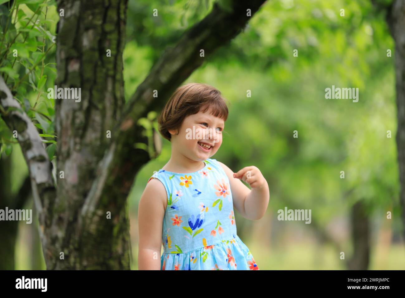 Una bambina carina che gioca sotto gli alberi da frutto, vestita con abiti estivi. Alberi verdi sfocati sullo sfondo. Giocare sotto gli alberi da frutto e mangiare frutta Foto Stock