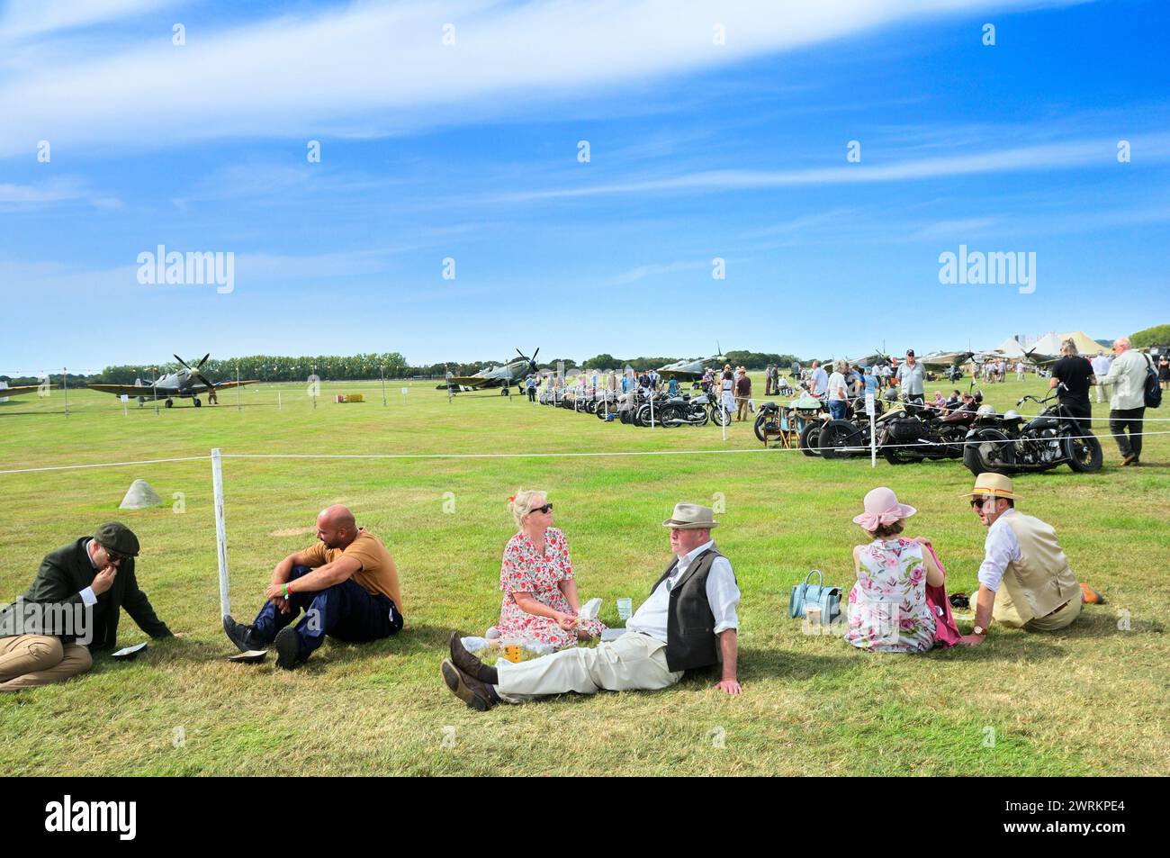 Persone sedute a rilassarsi sul campo d'aviazione in erba soleggiata vestite con abiti retrò al Goodwood Revival con motociclette classiche e aerei d'epoca, Regno Unito Foto Stock