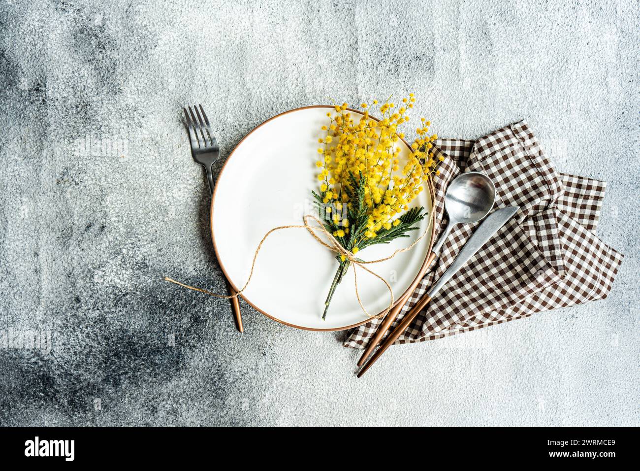 Vista dall'alto del tavolo rustico-chic con un delicato bouquet di fiori di mimosa legato a spago, completato da eleganti posate e un pannolino a scacchi Foto Stock