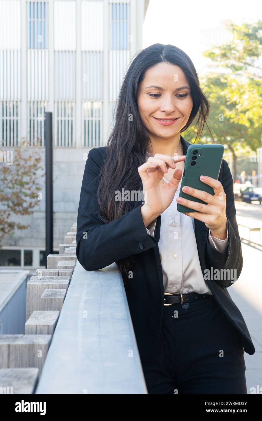 Una donna professionista in una tuta da lavoro impegnata con il suo smartphone all'esterno di un moderno edificio per uffici. Foto Stock