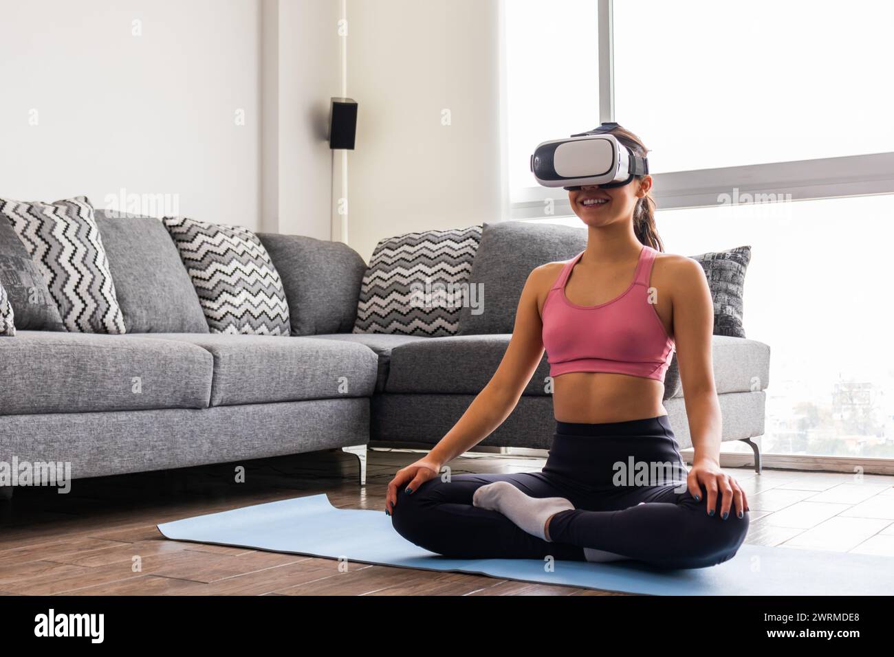 Una giovane donna adulta di etnia asiatica sorride mentre si siede a Sukhasana (posa facile), indossando un visore per la realtà virtuale in un soggiorno ben illuminato Foto Stock