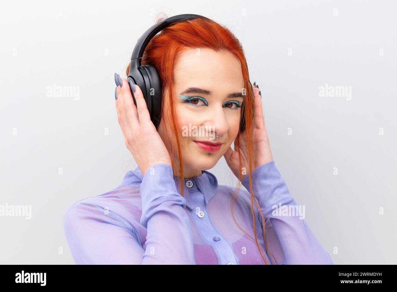 Una giovane donna d'affari adulta sicura di sé, con i capelli rossi e un piercing sul labbro inferiore, ascolta la musica con le cuffie guardando la fotocamera in piedi su una W bianca Foto Stock