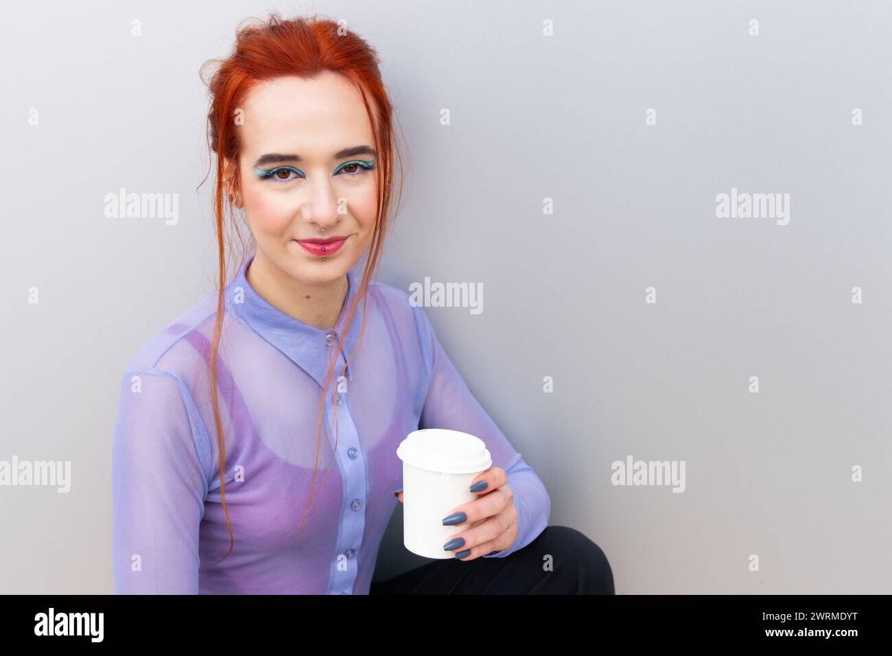 Una giovane donna d'affari trucco blu che mette in risalto gli occhi tenendo in mano una tazza di caffè da asporto bianca con una camicetta lilla trasparente che guarda la fotocamera Foto Stock