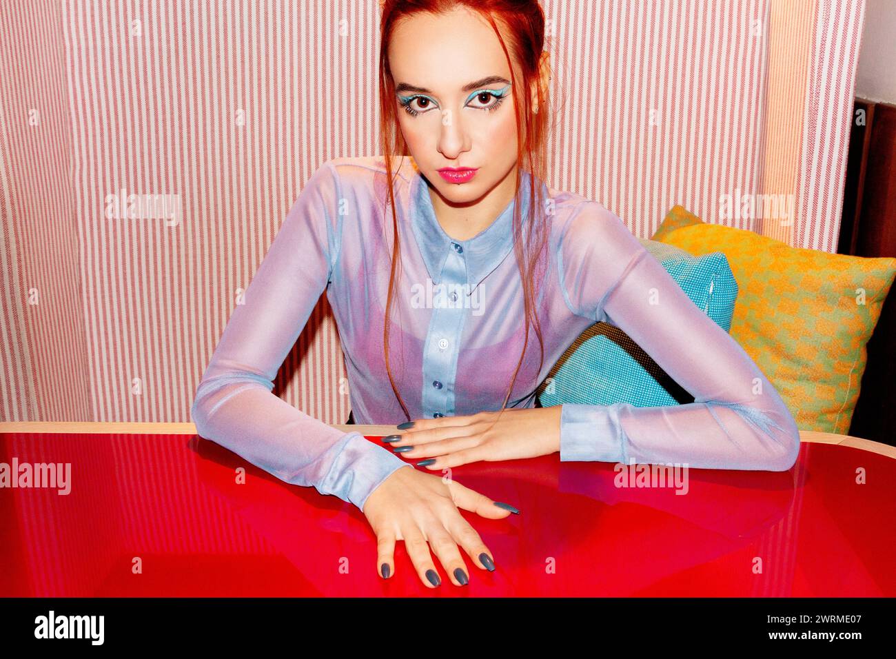 Una giovane donna caucasica che guarda la fotocamera con i capelli rossi tirati indietro, completando il suo audace trucco con un'ombra turchese e labbra rosa contro Foto Stock