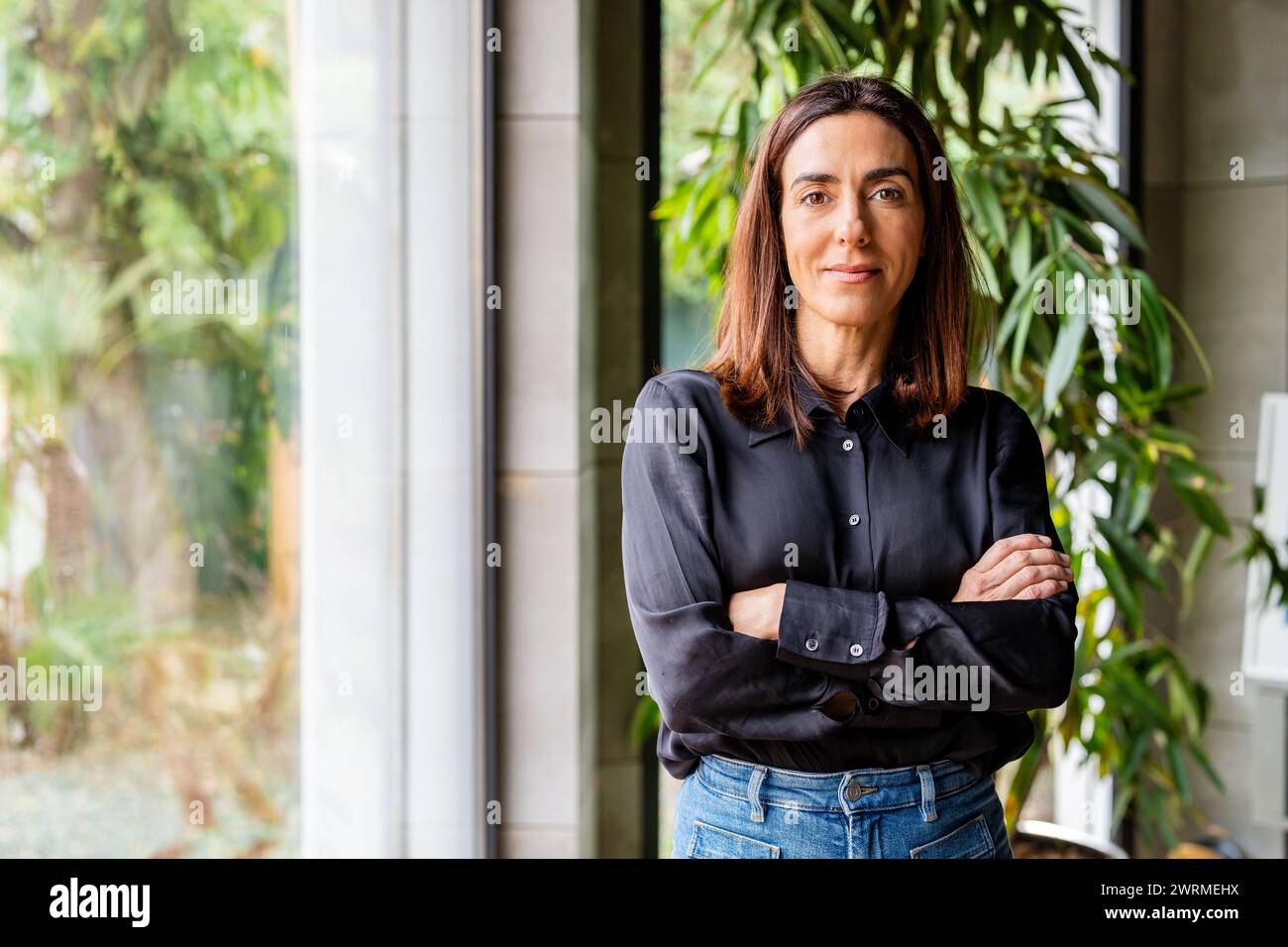 Una donna professionale con una postura sicura si trova in un ufficio ben illuminato, circondato da piante, che esemplifica leadership e competenza. Foto Stock
