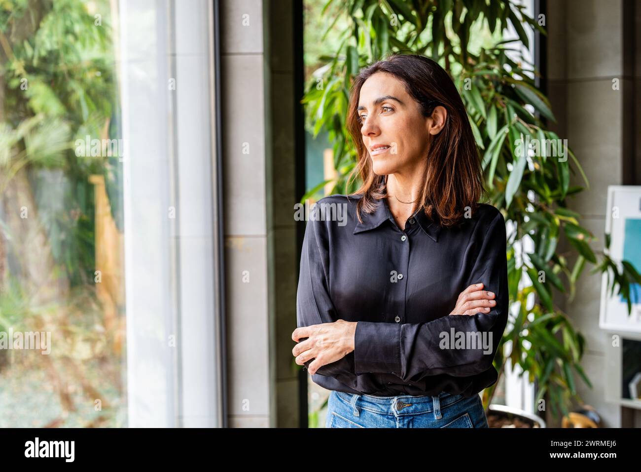 Imprenditrice donna che guarda lontano con fiducia, le braccia incrociate, in un ufficio contemporaneo con piante. Foto Stock