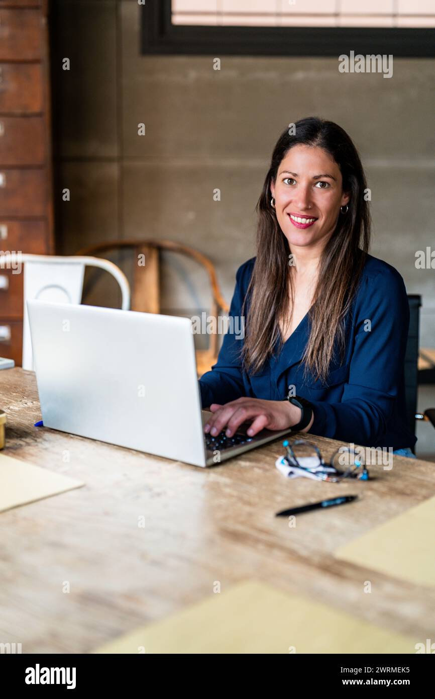 Una donna imprenditrice sicura di sé sta lavorando al suo laptop su una scrivania di legno, guardando la fotocamera con un sorriso amichevole. Foto Stock