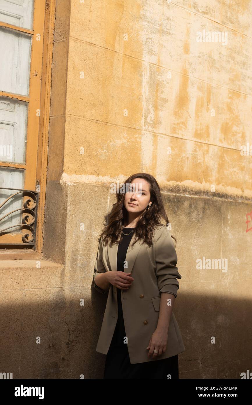 Una donna sicura di sé con un cappotto alla moda poggia su una parete intemprata con fascino rustico e un accenno di degrado urbano Foto Stock