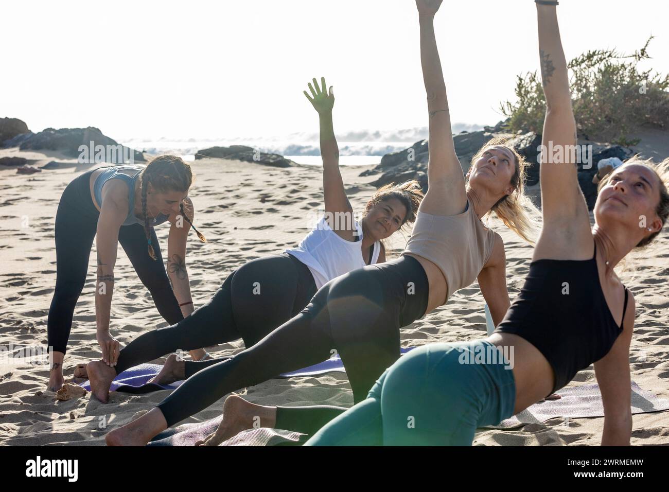 Un gruppo di donne partecipa a una lezione di yoga sulla spiaggia durante il tramonto, con una persona che esegue una posa Trikonasana. Foto Stock