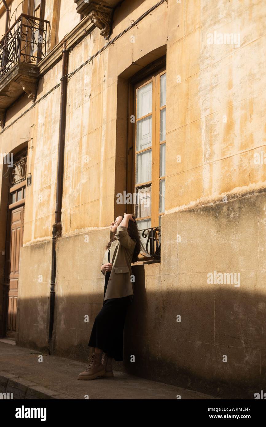 Una giovane donna si perde nel pensiero, la mano si tocca delicatamente i capelli, nel tranquillo calore di un vicolo storico illuminato dal sole Foto Stock