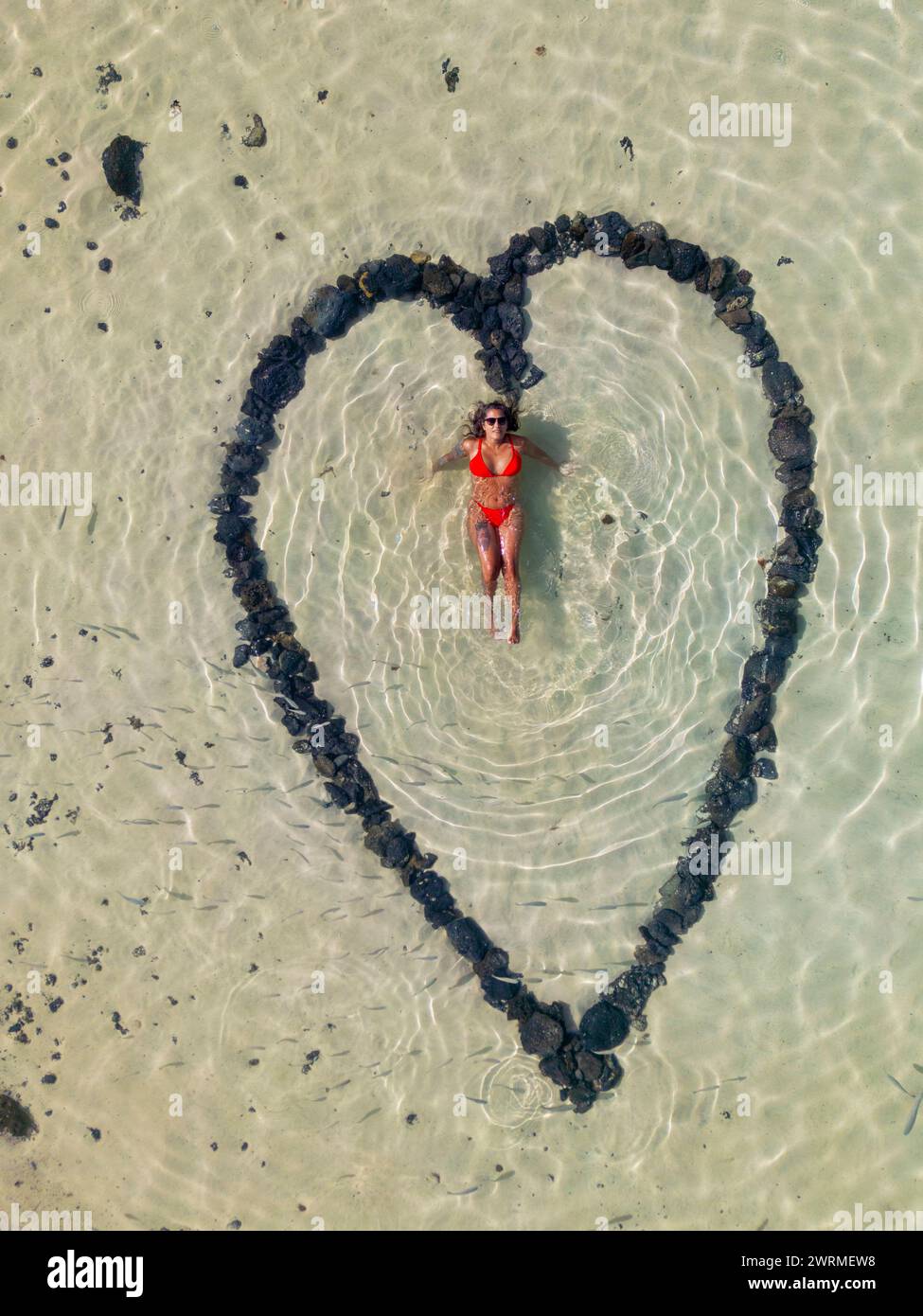 Vista aerea di una formazione a forma di cuore fatta di pietre su una spiaggia sabbiosa con una donna sdraiata al centro, catturando l'essenza dell'amore estivo. Foto Stock