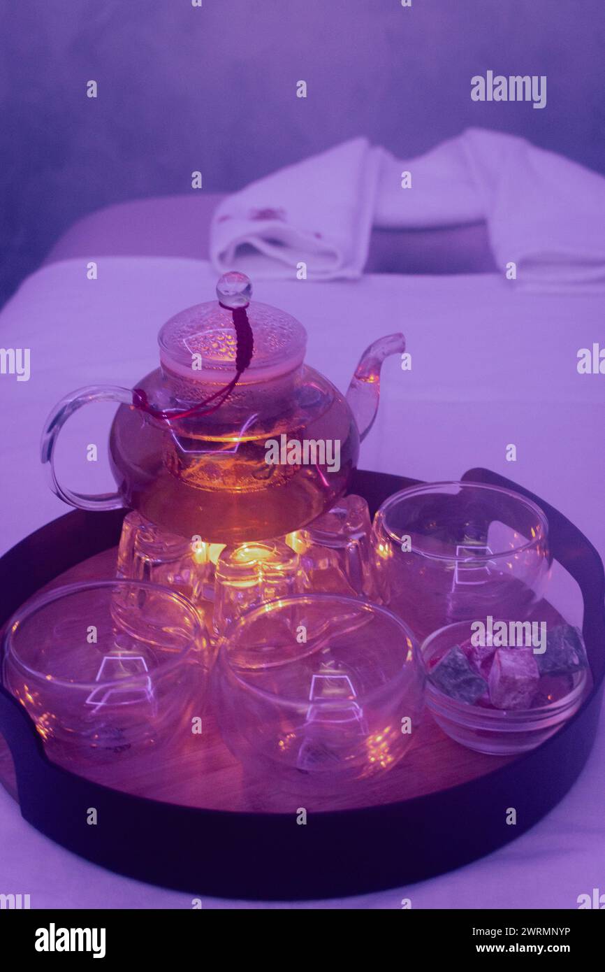 Teiera di vetro con tè alle erbe e candela sul divano per massaggi. Interno del salone di bellezza. Stile di vita di lusso. Centro termico. Teiera con tazze sul vassoio Foto Stock
