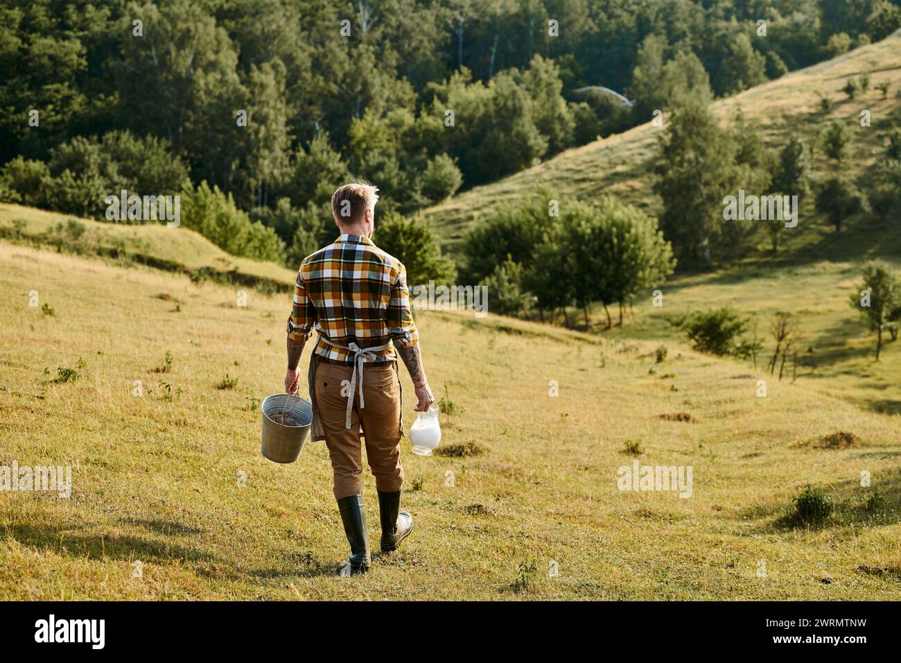 vista posteriore di un contadino moderno e laborioso con tatuaggi che camminano con secchio e vaso di latte in mano Foto Stock