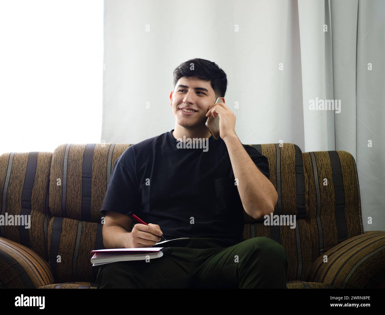 Immagine di un giovane sorridente che parla allegramente al telefono, seduto sul divano a casa e prende appunti dei dati che gli sono passati durante la chiamata Foto Stock