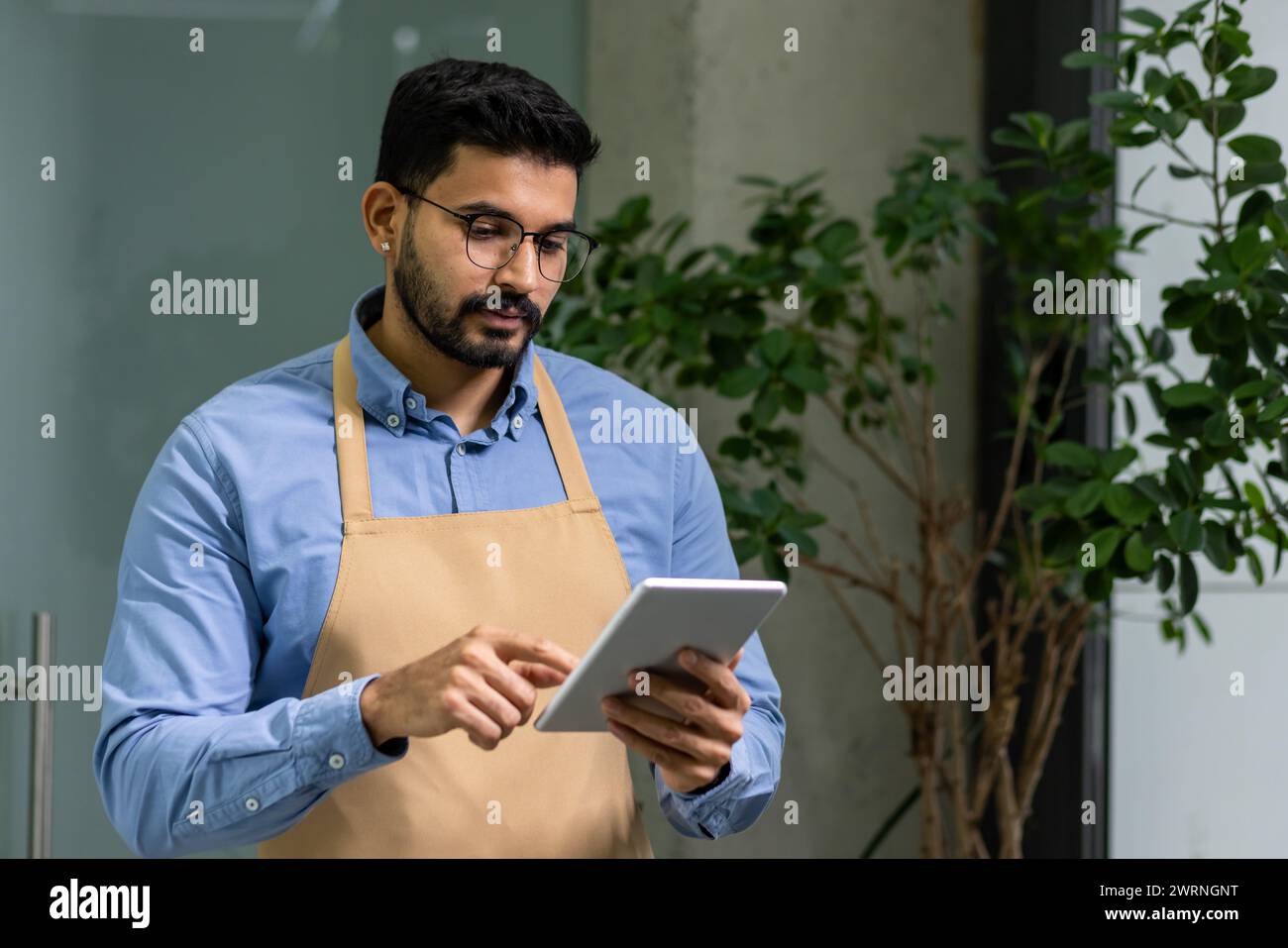 Un uomo dedicato con barba e occhiali si concentra sul tablet indossando un grembiule casual, incarnando l'imprenditorialità e la cura delle piante. Foto Stock